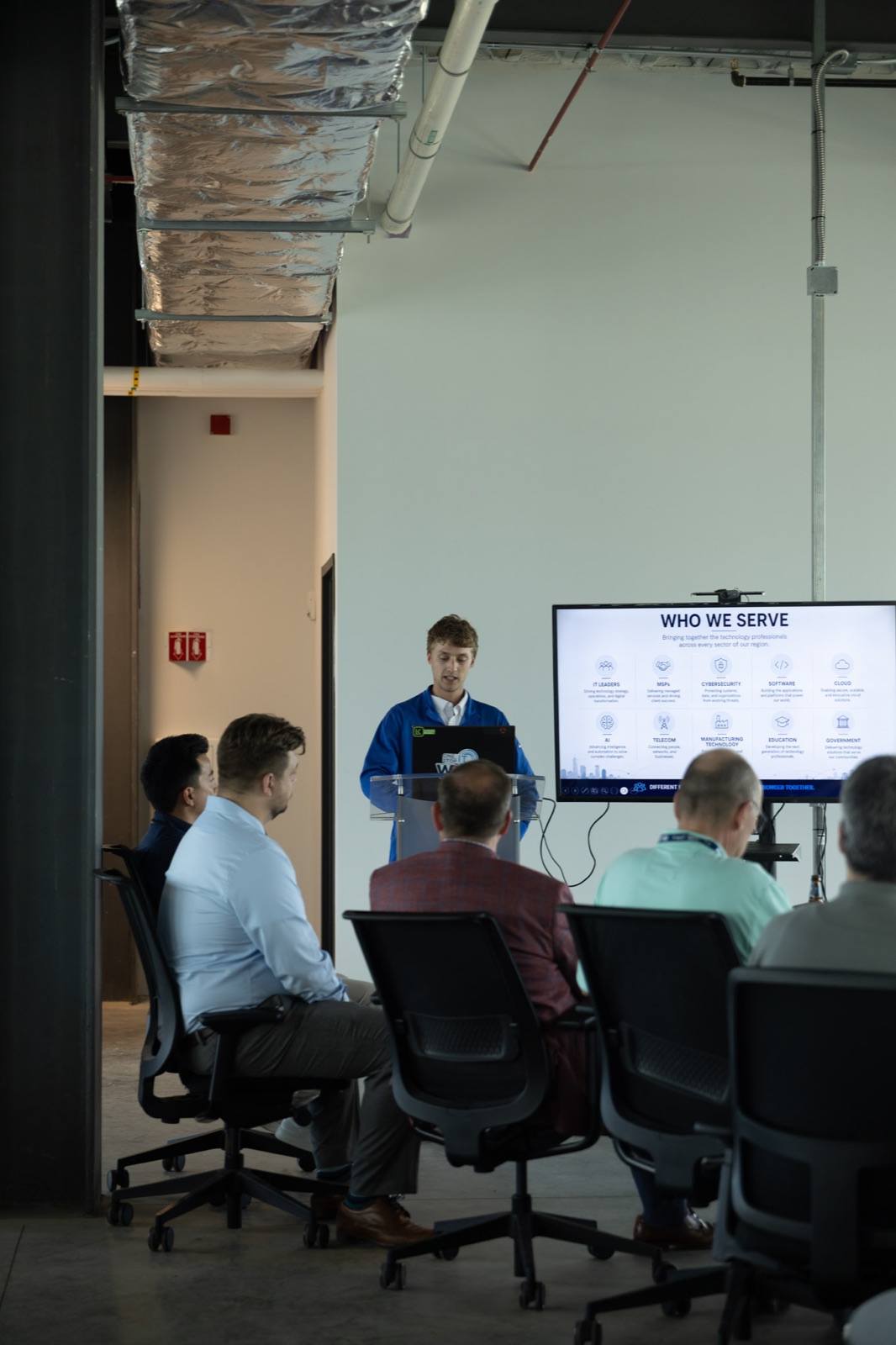 A presenter addressing attendees beside a screen describing who Great Lakes Technology Corridor serves.